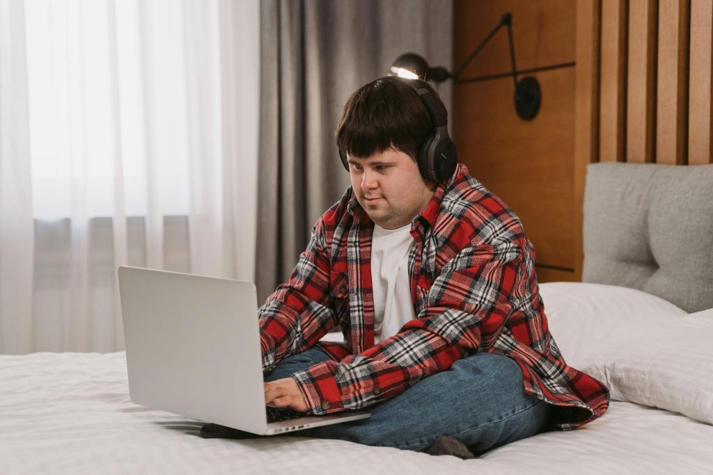 A young man with headphones uses a laptop while sitting on a bed, embracing a relaxed lifestyle indoors.